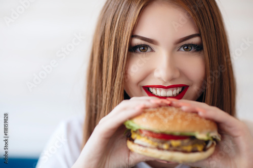 Beautiful woman with red long straight hair bright make-up brown eyes red lipstick long eyelashes pink nail polish in the hands holding a large hamburger  sitting at a table in a cafe dinner alone