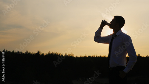 Young businessman drinking alcohol at sunset, silhouette against the sky