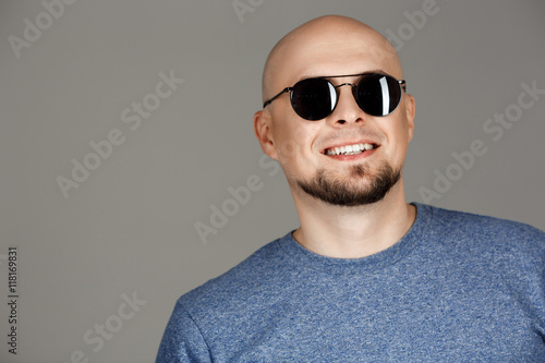 Portrait of confident handsome middle-aged man in grey shirt and sunglasses posing smiling at camera over dark background.