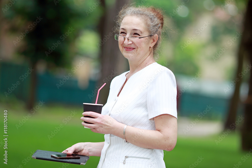 женщина в возрасте с гаджетом в руках пьет кофе в парке