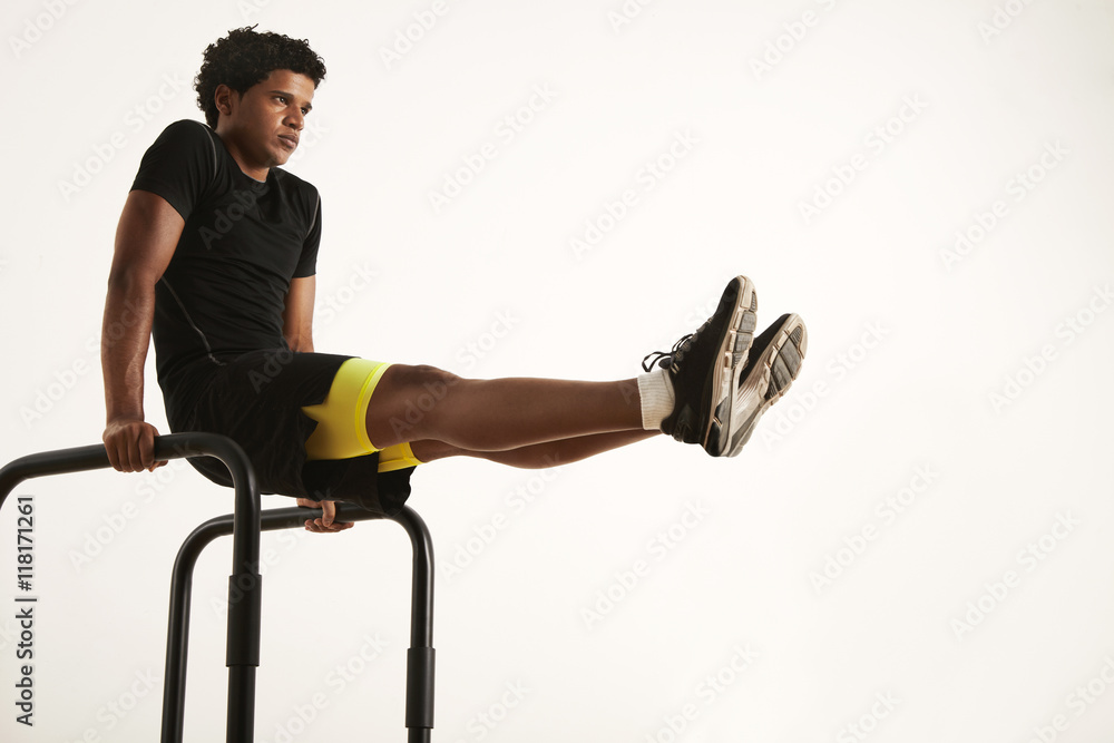 Obraz premium Serious young fit African American man in black sportswear doing L-sits on short bars at home against a white wall