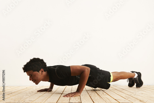 Low angle shot of a smiling fit young black athlete doing pushups on a light wooden floor against a white wall
