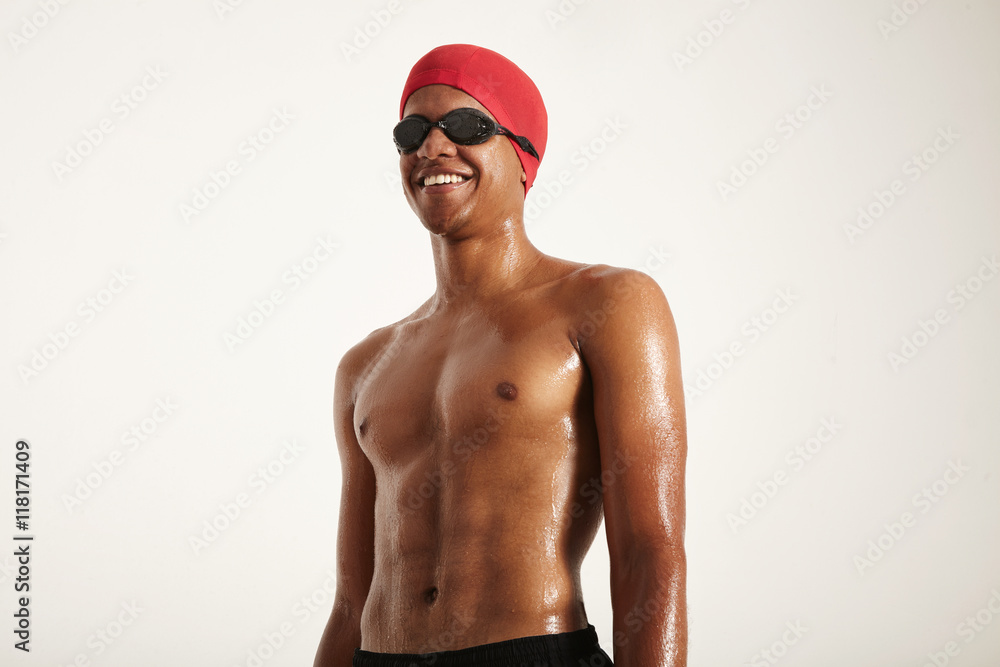 Low angle shot of a happy fit muscular smiling African American swimmer with wet skin wearing red cap and black goggles looking away from the camera isolated on white. Low angle shot of a happy fit muscular smiling African American swimmer with wet skin wearing red cap and black goggles looking away from the camera isolated on white.