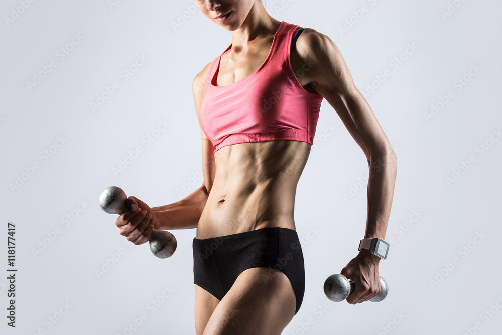 Fototapeta premium Attractive young fitness person wearing smartwatch and red sportswear top lifting dumbbells. Serious sporty model girl working out, doing weight training with dumbbells on grey background. Close-up