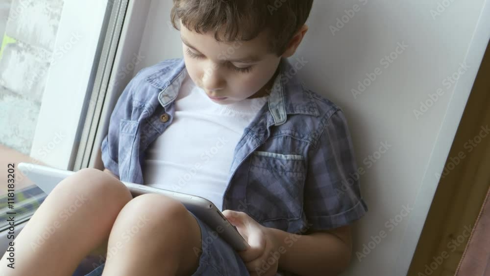PORTRAIT: A cute little boy uses a white tablet PC on a windowsill at home PORTRAIT: A cute little boy uses a white tablet PC on a windowsill at home
