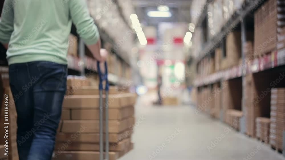 A man is pushing a trolley full of boxes on it between palettes with cardboard boxes in a storage warehouse A man is pushing a trolley full of boxes on it between palettes with cardboard boxes in a storage warehouse
