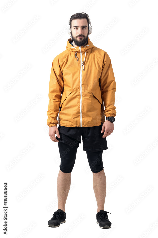 Young bearded jogger in sportswear wearing ocker jersey jacket with headphones looking at camera. Full body length portrait isolated on white background.