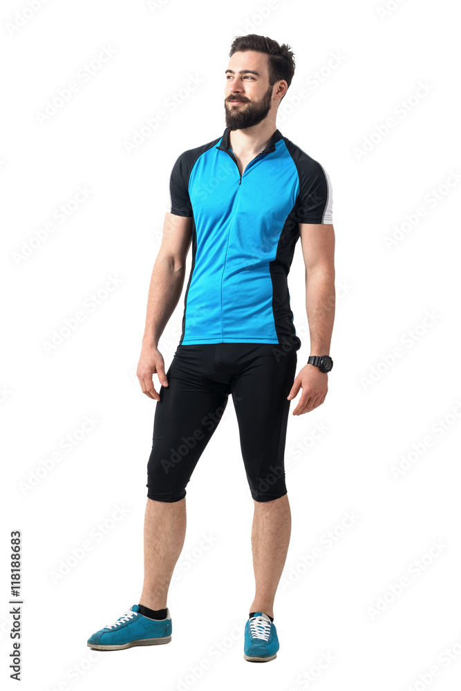 Young bearded cyclist guy in blue jersey t-shirt and leggings relaxed smiling and looking away. Full body length portrait isolated over white studio background. Young bearded cyclist guy in blue jersey t-shirt and leggings relaxed smiling and looking away. Full body length portrait isolated over white studio background.
