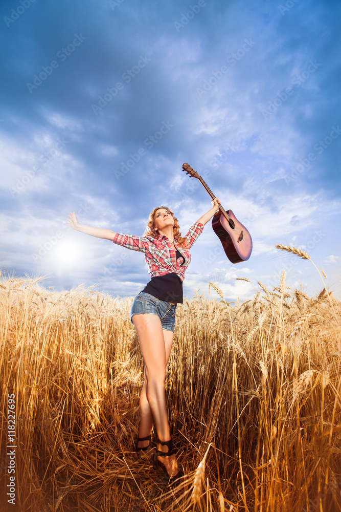 Girl in plaid shirt and jeans shorts walking in golden wheat field with acoustic Guitar. young adult caucasian girl walk outdoor against evening sky. Summer season. Horizontal Girl in plaid shirt and jeans shorts walking in golden wheat field with acoustic Guitar. young adult caucasian girl walk outdoor against evening sky. Summer season. Horizontal