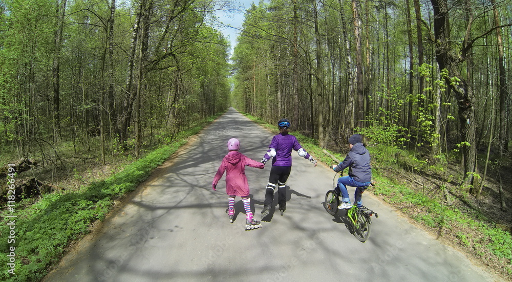 Fototapeta premium Mother with children ride on rollerblades and bike by road with bicyclists at sunny spring day in forest. Aerial view