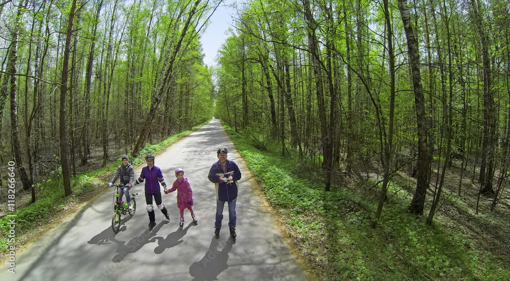 Four people parents and children ride on rollerblades and bicycle by road along forest at sunny spring day. Aerial view Four people parents and children ride on rollerblades and bicycle by road along forest at sunny spring day. Aerial view