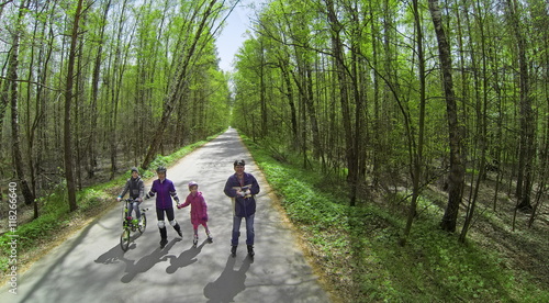 Four people parents and children ride on rollerblades and bicycle by road along forest at sunny spring day. Aerial view