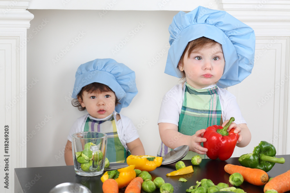Little girl in kitchen apron and cap with large red pepper and small boy Little girl in kitchen apron and cap with large red pepper and small boy