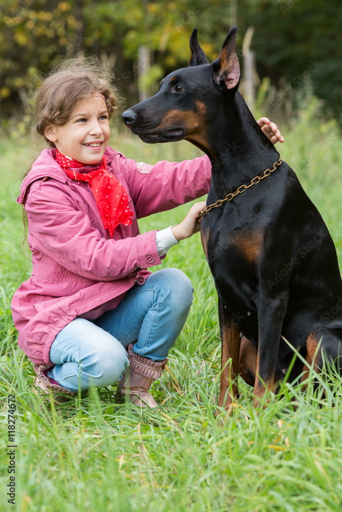 Little curly girl with red scarf is holding big black dog on the chain in park. Little curly girl with red scarf is holding big black dog on the chain in park.