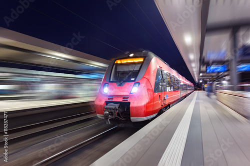Modern high speed red passenger train moving through railway station at night. Railway station at sunset in Nuremberg, Germany. Railroad with motion blur effect. Industrial concept landscape