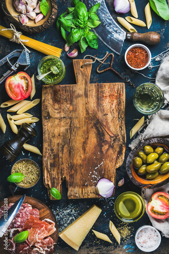 Italian food cooking ingredients on dark background with rustic wooden chopping board in center, top view, copy space, vertical composition