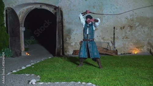 A young man is swinging his whip as a part of the performance. He is also having one hand on his ear. Wide-angle shot. It is evening time.
