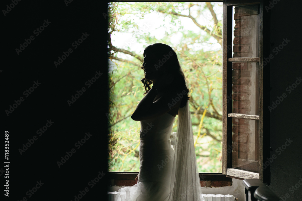 Bride with a tender figure stands in the front of an open window