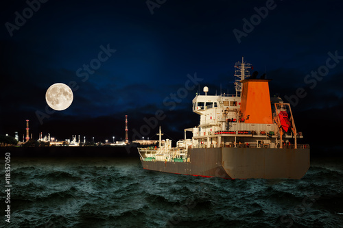 Tanker ship on sea with oil refinery industry in background at night.