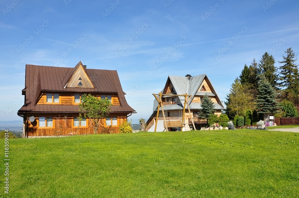Two old houses with pitched roof in Zakopane style on a background of green grass and blue sky. Two old houses with pitched roof in Zakopane style on a background of green grass and blue sky.