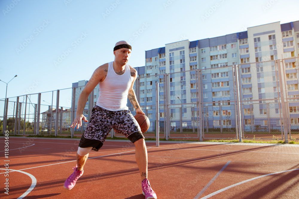 Obraz premium Guy plays basketball on a red Court in sunny weather the background of houses. Solar flare in the frame.