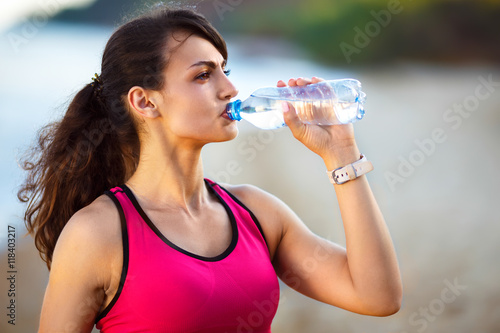 Fitness beautiful woman drinking water and sweating after exercising on summer hot day in beach. Female athlete after work out.

