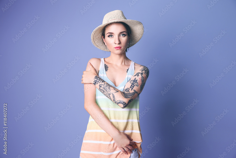 Beautiful young woman with tattoo wearing hat and posing on blue background