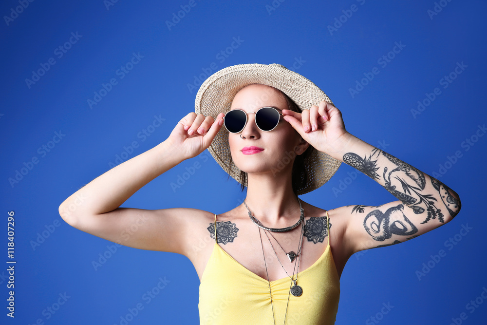 Beautiful young woman with tattoo wearing hat and sunglasses on blue background Beautiful young woman with tattoo wearing hat and sunglasses on blue background