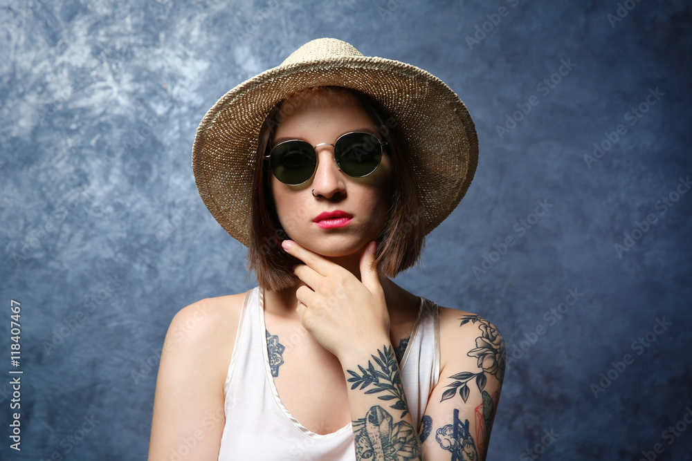 Beautiful young woman with tattoo wearing sunglasses and hat on gray background Beautiful young woman with tattoo wearing sunglasses and hat on gray background