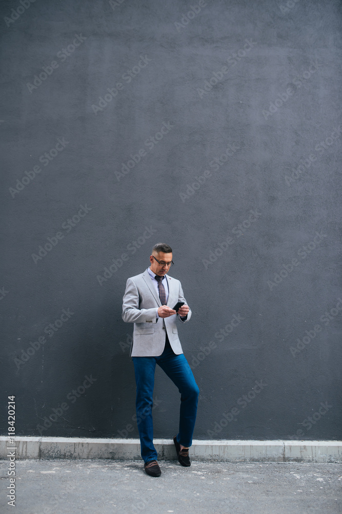Shot of handsome mature businessman typing massage while standing near grey wall. Shot of handsome mature businessman typing massage while standing near grey wall.