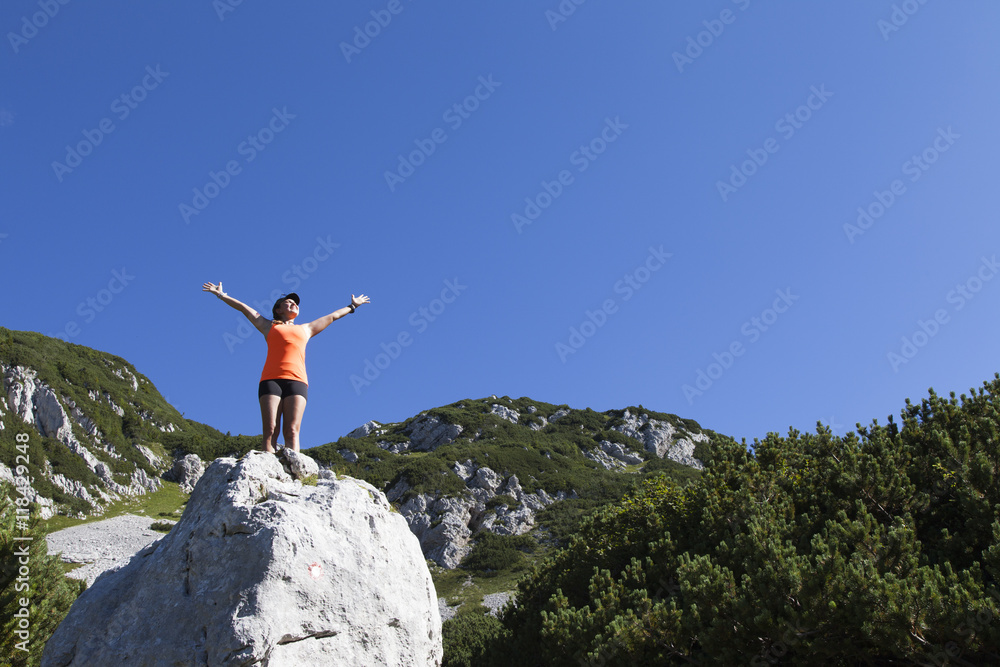 Obraz premium Young pretty smiling woman hiker standing on a rock with raised outstretched hands, high in the mountains, freedom and happiness, achievement in mountains 