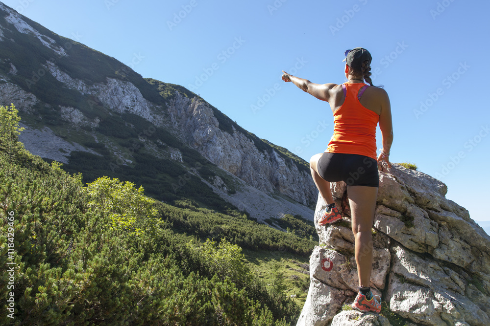 Young woman hiker points on mountain peak in the Alps, on sunny day, against clear blue sky, selective focus, space for text 