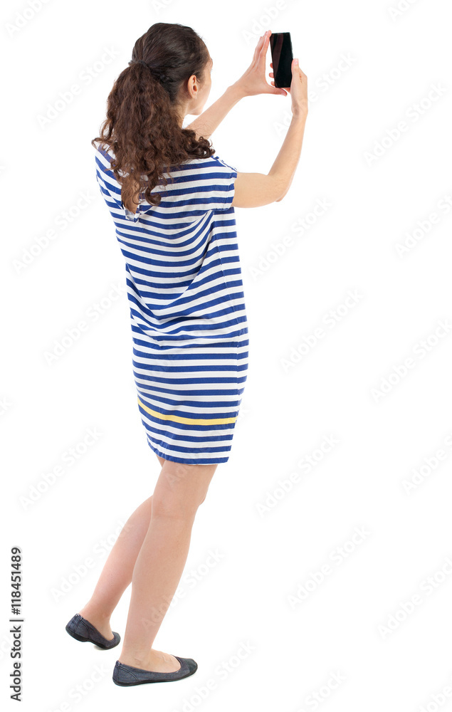 Fototapeta premium back view of standing young beautiful woman using a mobile phone. girl watching. Rear view people collection. backside view of person. Isolated over white background. Swarthy girl in a checkered
