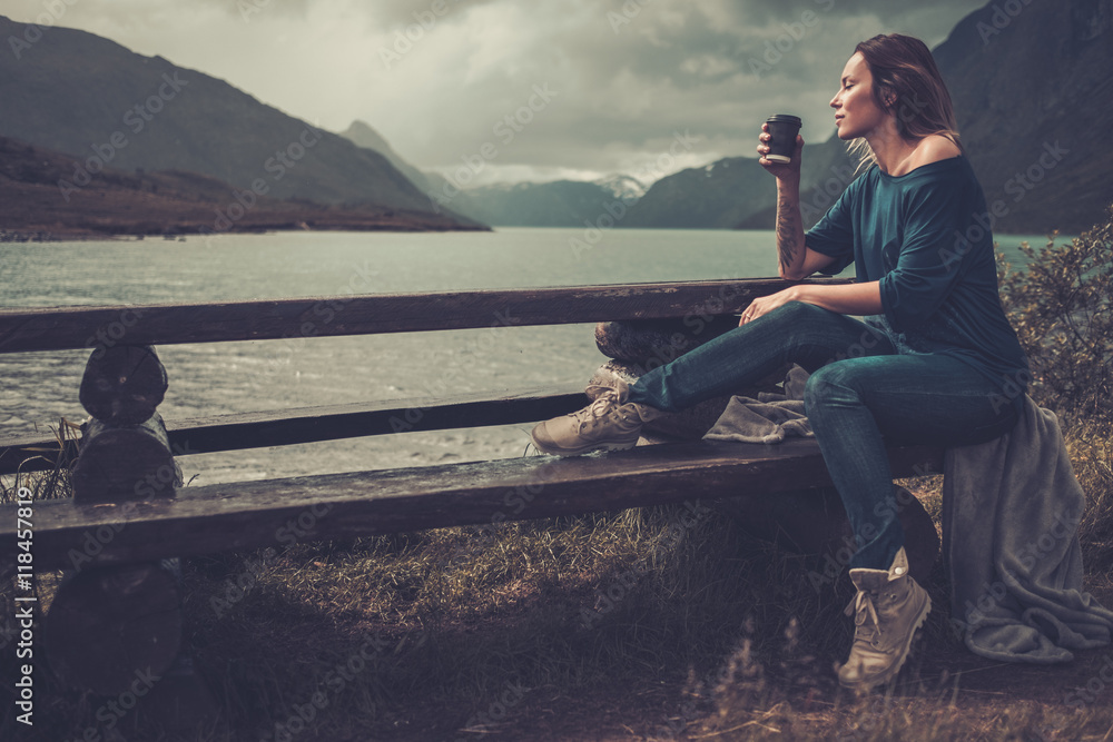 Beautiful woman with cup of coffee or tea sitting on a banch near the wild lake, with mountains on the background.