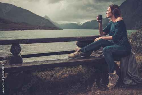 Beautiful woman with cup of coffee or tea sitting on a banch near the wild lake, with mountains on the background.