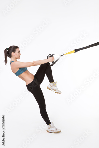 Attractive young woman exercising with modern sports equipment. Picture of fitness lady training with suspension trainer sling or suspension straps in studio.