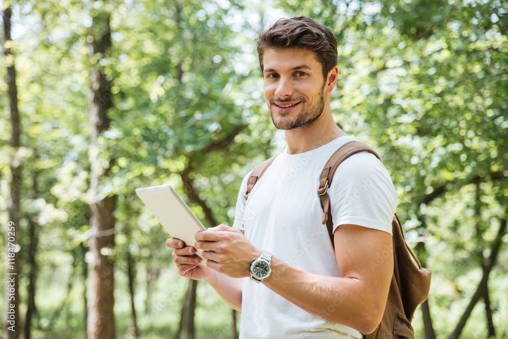 Cheerful young man with backpack using tablet in forest Cheerful young man with backpack using tablet in forest