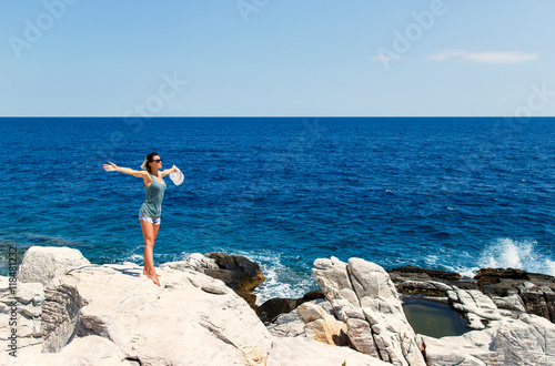 Young woman standing on the cliff against sea and blue sky, with her arms outstretched.