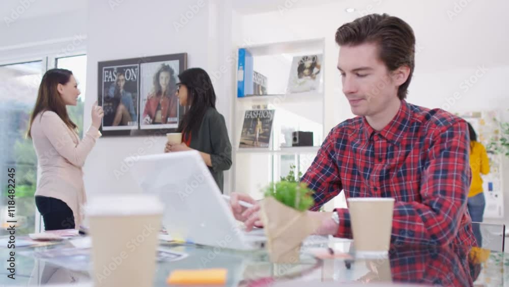 Worker in creative office, working on laptop with colleagues working behind Worker in creative office, working on laptop with colleagues working behind