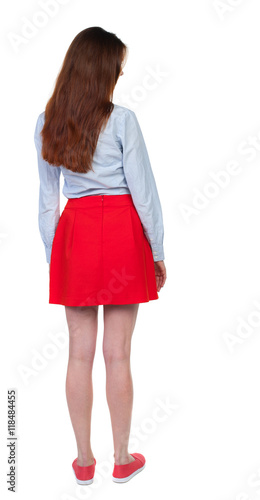 back view of standing young beautiful woman. girl watching. Rear view people collection. backside view of person. Long-haired brunette in red skirt looks down sideways. 