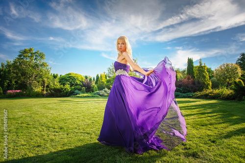 Blonde gorgeous woman in long purple luxurious dress posing in garden. Dress fluttering on wind.