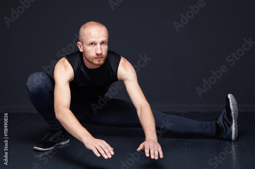 Young fitness man performing legs stretching against dark background