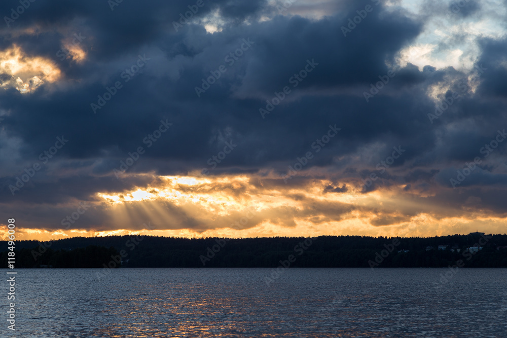 View of dark and dramatic clouds, rays of light and a lake in Finland at sunset in the summer.