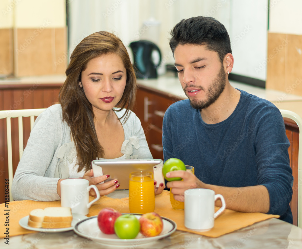 Young charming couple sitting by breakfast table looking at tablet screen, fruits, juice and coffee placed in front, hostel environment Young charming couple sitting by breakfast table looking at tablet screen, fruits, juice and coffee placed in front, hostel environment