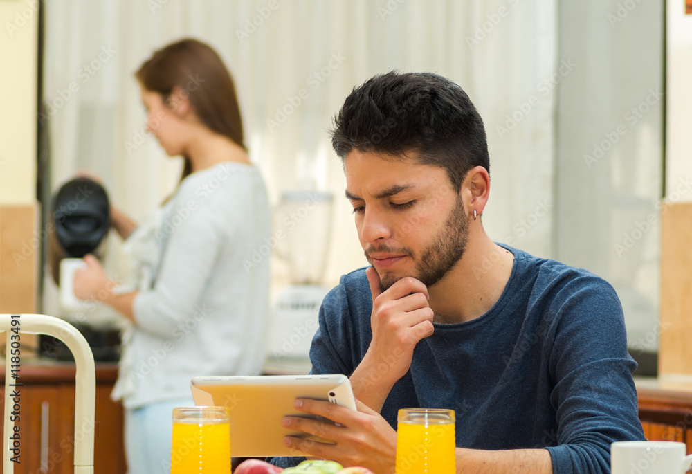 Young handsome man seated by breakfast table looking at tablet screen, girl standing behind pouring hot water, fruits, juice placed in front, hostel environment Young handsome man seated by breakfast table looking at tablet screen, girl standing behind pouring hot water, fruits, juice placed in front, hostel environment