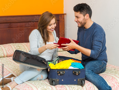 Young charming hispanic couple wearing casual clothes sitting on bed packing into suitcase together, hostel guest concept