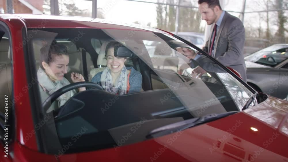 Salesman talking to women in car dealership & showing them new car Salesman talking to women in car dealership & showing them new car