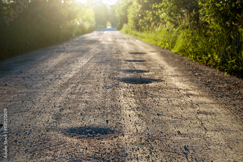 Bumpy dirt road with holes against sunshine in background. Shallow depth of field.