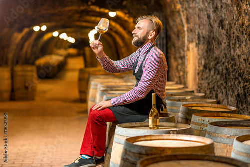 Handsome sommelier in apron and checkered shirt looking at wine glass sitting on the barrel in the old cellar.