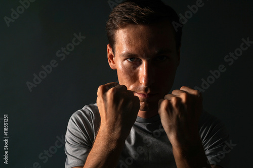 Young man in t-shirt on a dark background holding fists at his face.